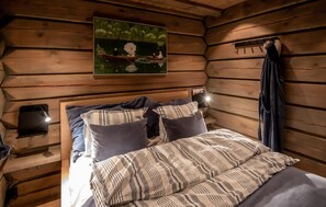Ein Zimmer einer Holzblockhütte mit einem Bett, Kissen, einem Bild eines Bootes auf dem Wasser und einem Nachttisch mit einer Lampe.