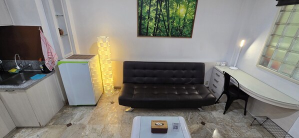 Cocina compacta con refrigerador, lavamanos y zona de comedor con sofá y una mesa pequeña.