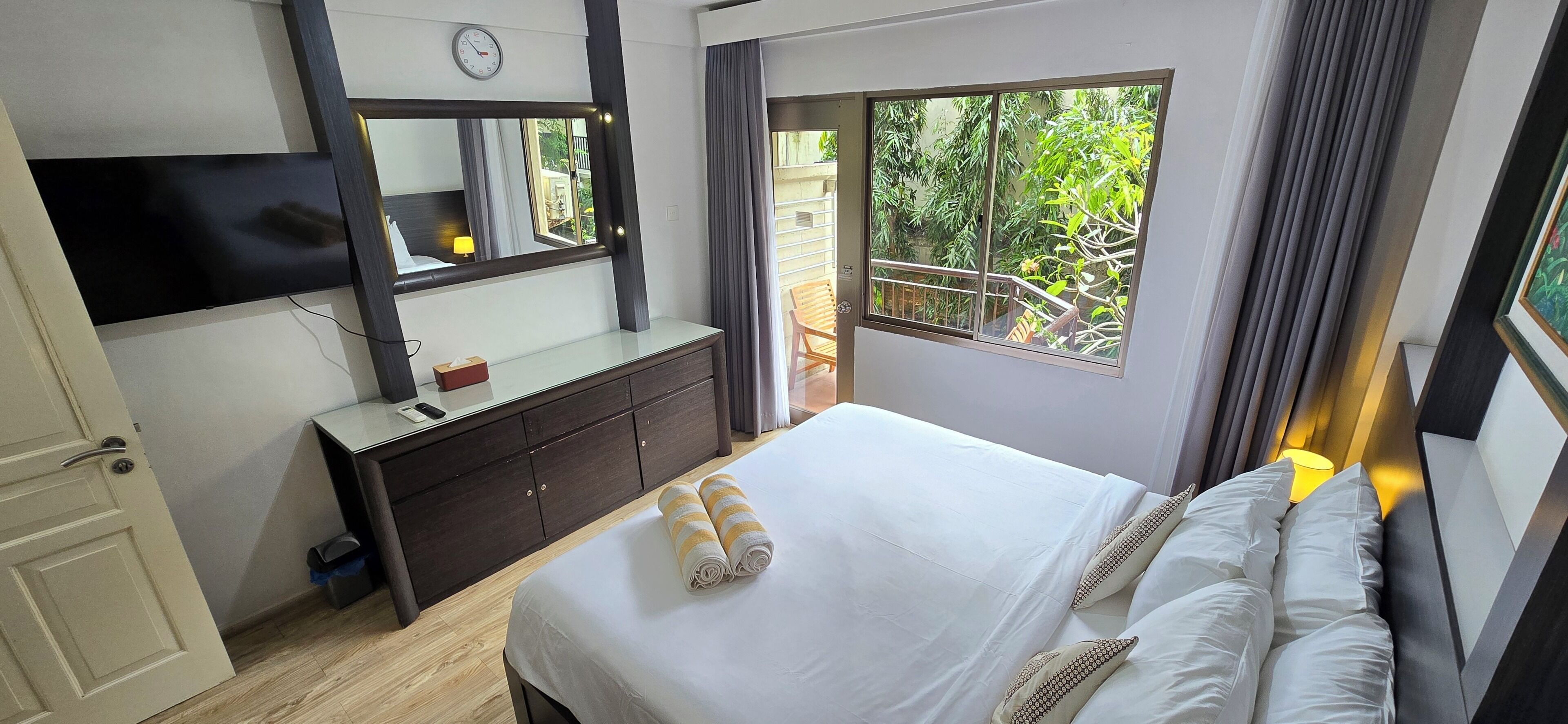 A hotel room with a bed, a dresser, a TV, a window with a view of greenery, and a clock on the wall.