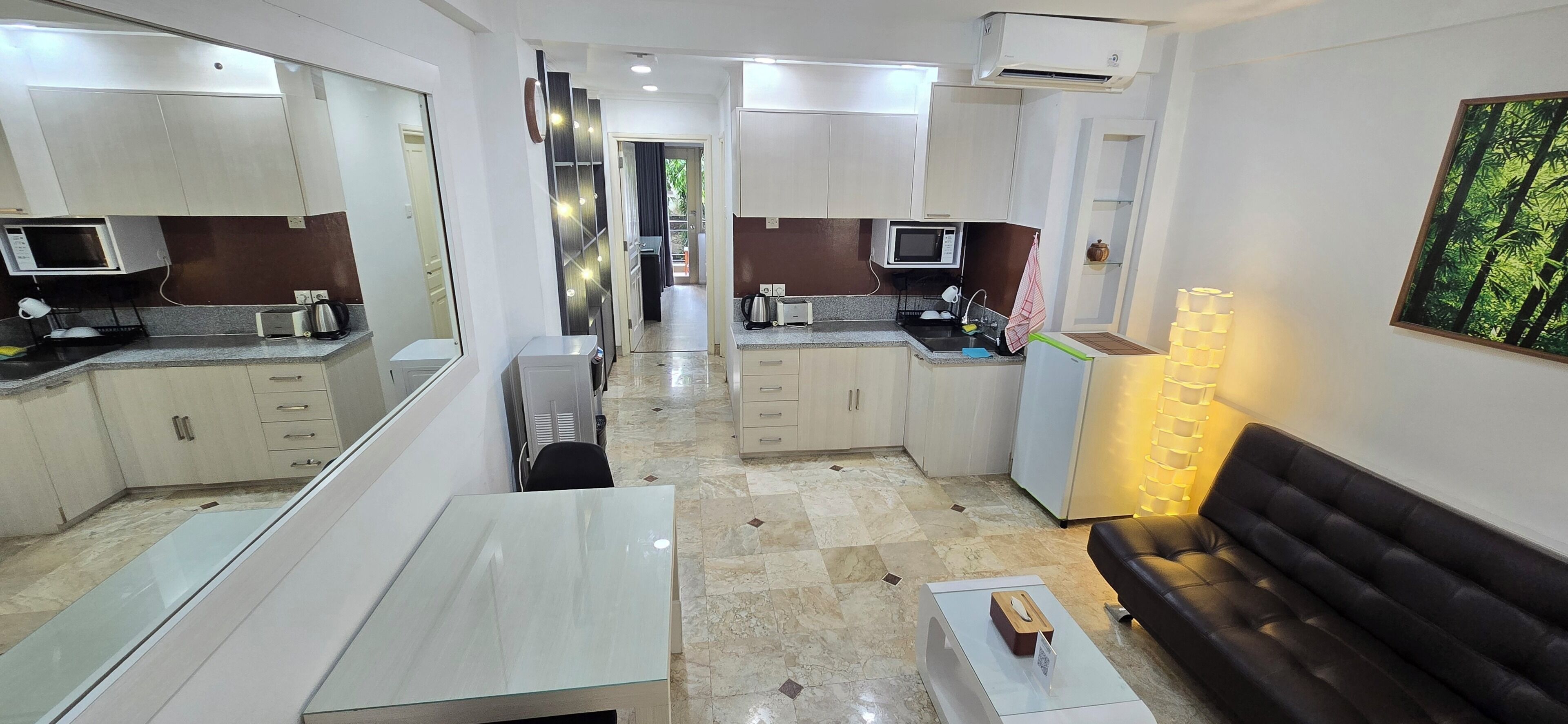 A modern kitchen with a microwave, toaster, and coffee maker. There is a dining area with a white table and a black sofa. The room has a large mirror and a painting of a forest.