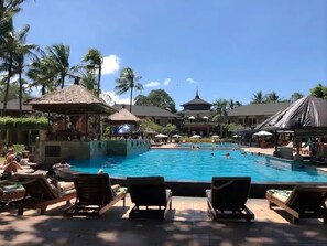 Área de piscina en un resort con sillones de descanso, una piscina central y un edificio con techo abovedado.