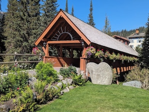 A wooden cabin with a sloped roof, surrounded by a garden with various plants and flowers, and a large rock in the foreground. - Cozy Alpine Condo w/Hot Tub! (Sun Peaks)