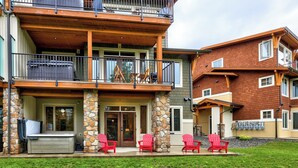 A two-story building with a stone facade, a balcony with a metal railing, and a set of red Adirondack chairs in front. - Cozy Alpine Condo w/Hot Tub! (Sun Peaks)