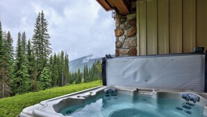 A hot tub with a stone wall backdrop, surrounded by a forest and a mountain view. - Cozy Alpine Condo w/Hot Tub! (Sun Peaks)