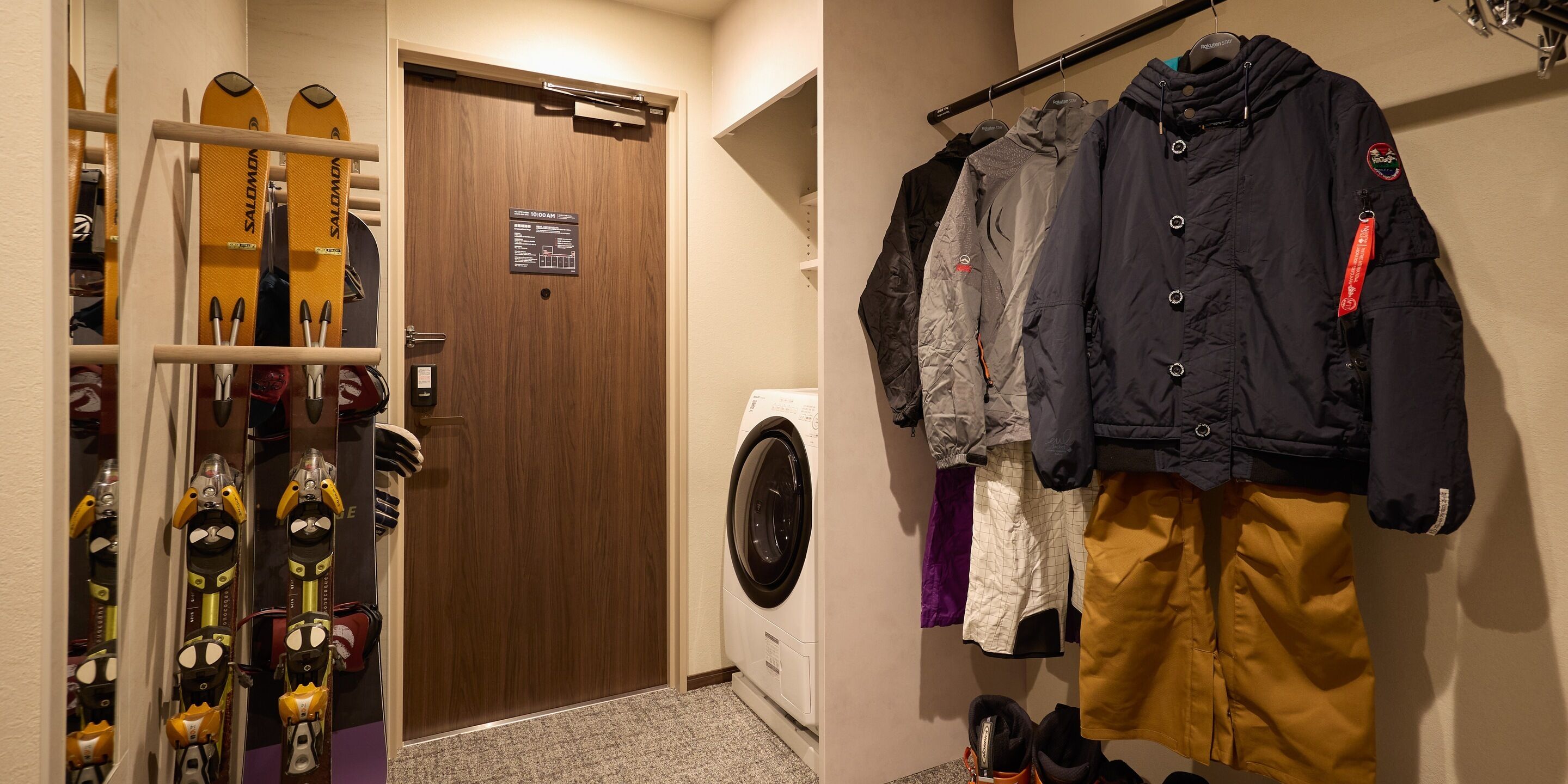 A hotel room with a wooden door, a washing machine, ski equipment, and a rack with jackets and pants.