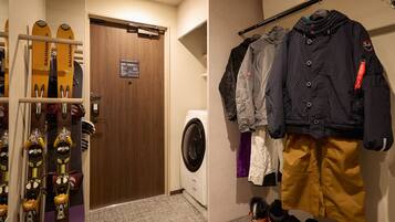 Habitación de hotel con puerta de madera, lavadora, equipo de esquí y un perchero con chaquetas y pantalones.