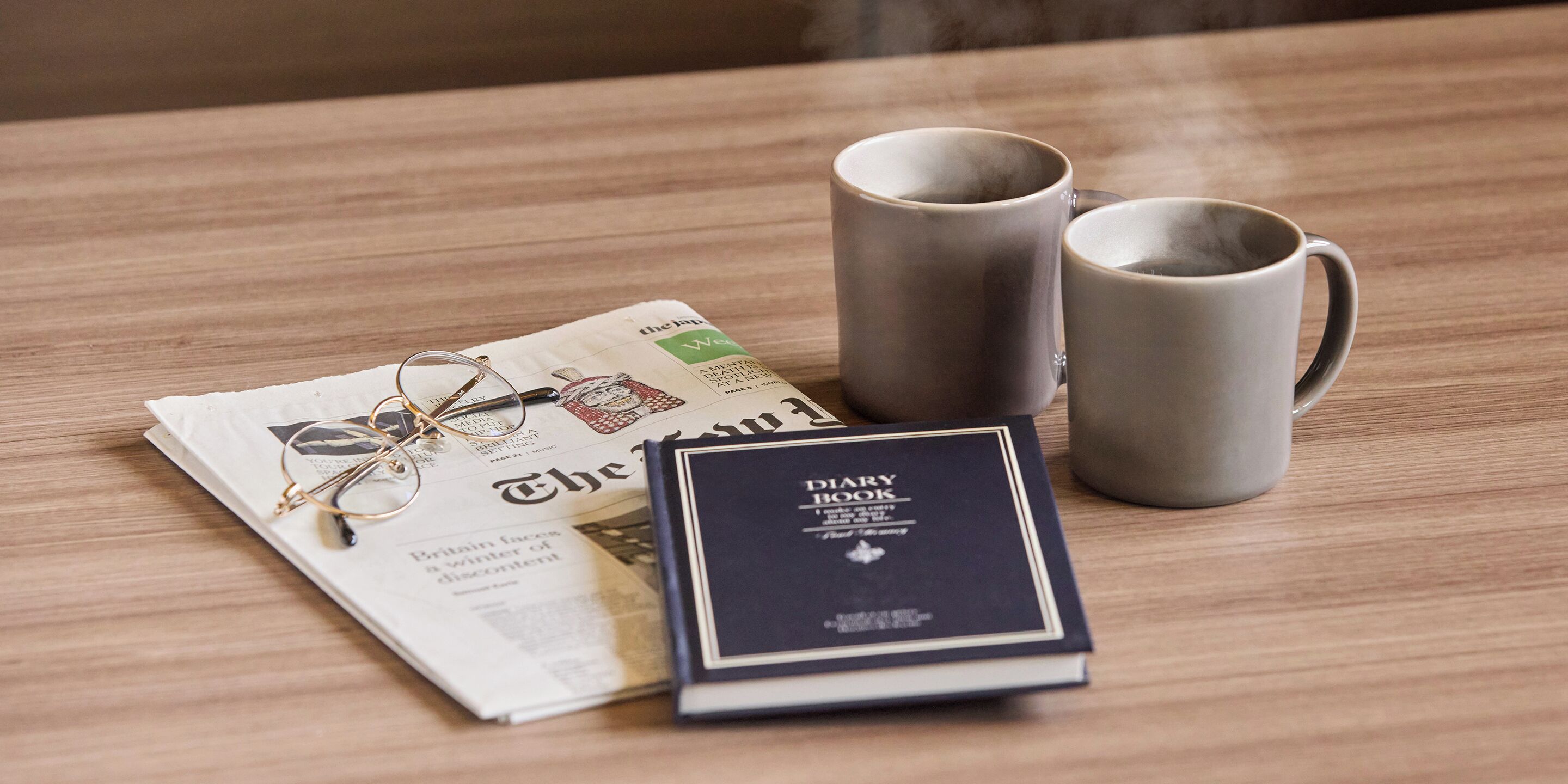 A wooden table with a pair of glasses, a diary, and two mugs.