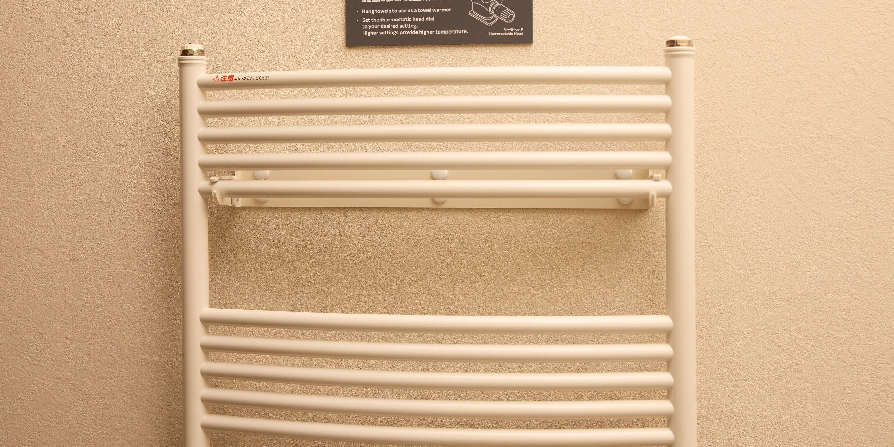 Een witte handdoekwarmer met een thermostaatknop en een waarschuwingstickker in een badkamer.