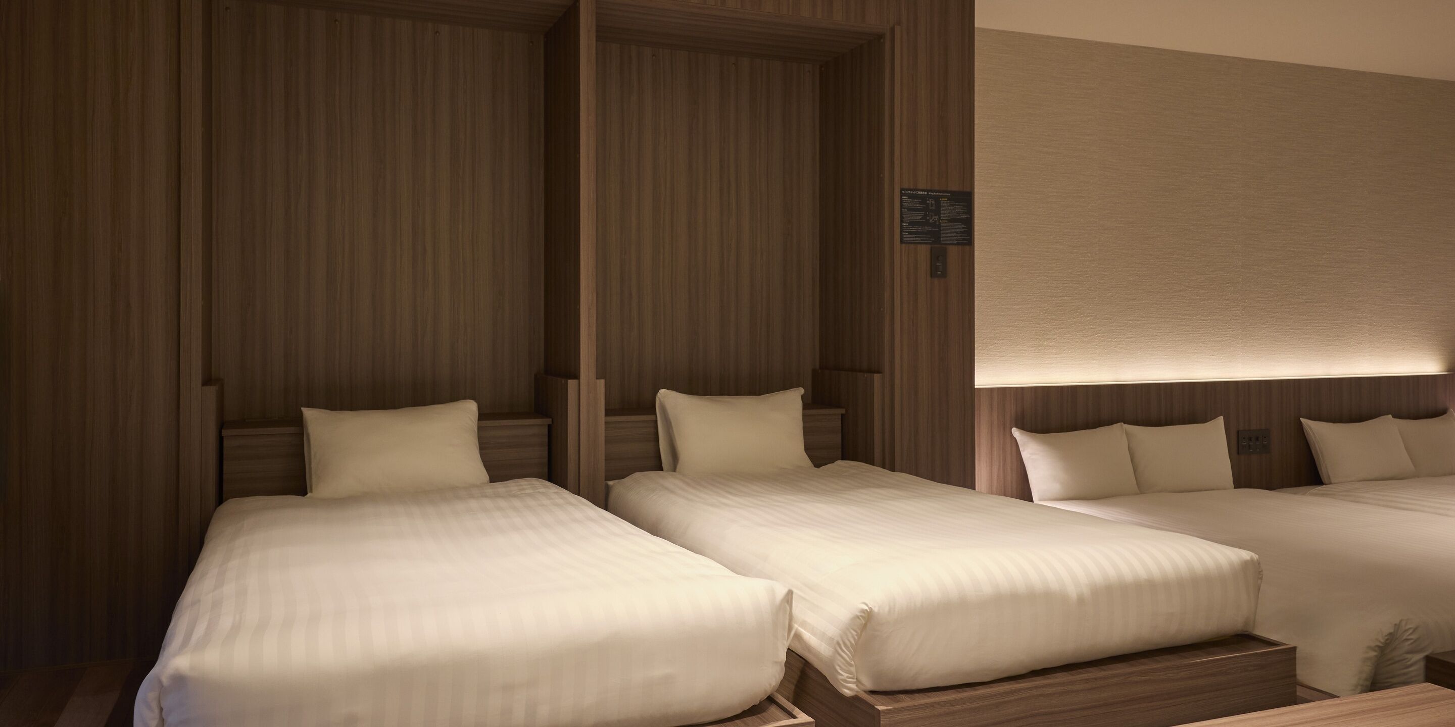 Een hotelkamer met drie bedden op een rij, houten lambrisering aan de wanden en een lamp boven de bedden.