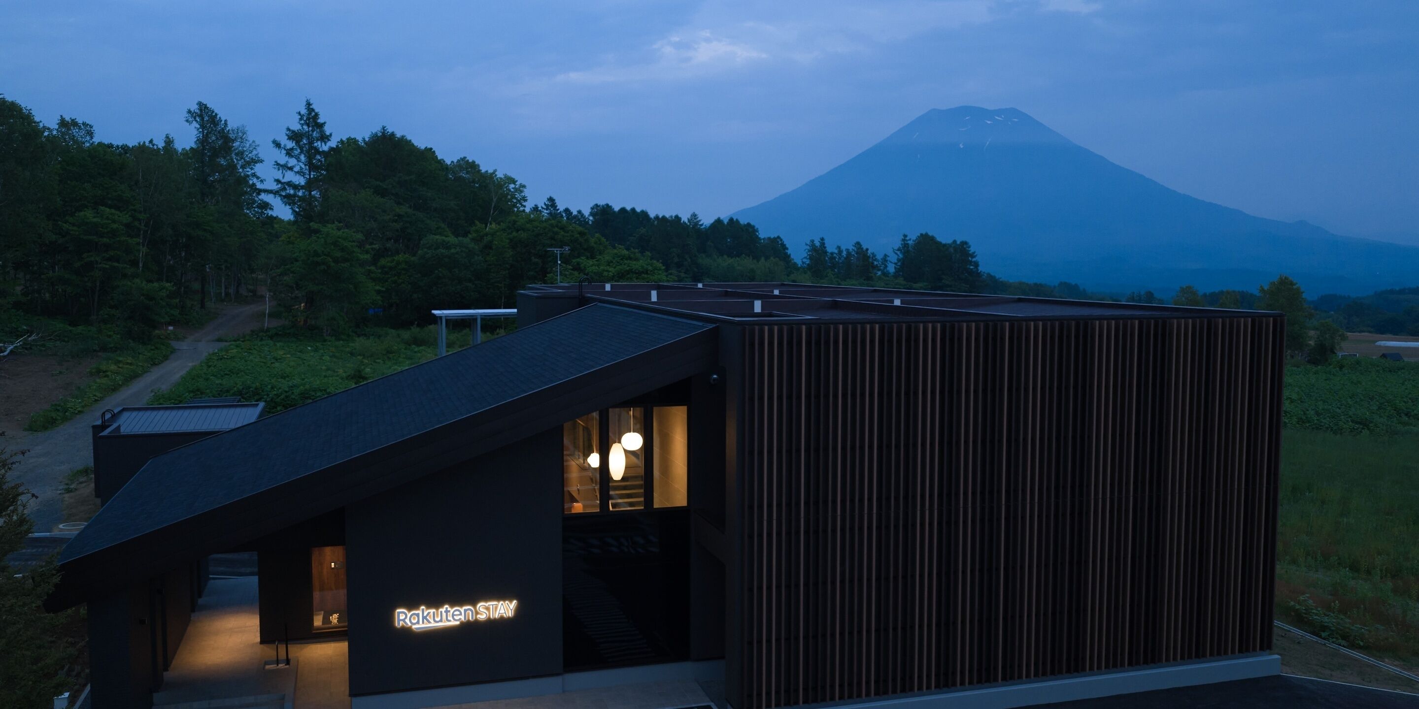 Un bâtiment moderne doté d’un toit en pente, d’une entrée bien visible et d’une enseigne indiquant « Rakuten STAY ».