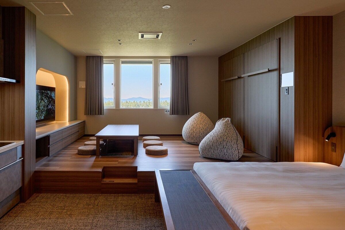 A modern hotel room with a bed, a desk, a chair, a sofa, and a view of the outdoors.