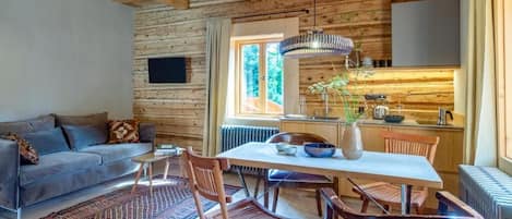 A cozy cabin interior with a wooden ceiling, a dining area with a table and chairs, a sofa, and a kitchen area.