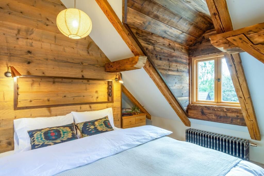 A bedroom with a sloped ceiling, wooden beams, a bed with patterned pillows, a bedside table, and a window.