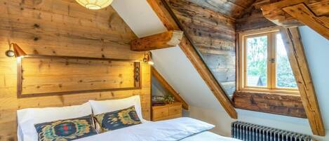 A bedroom with a sloped ceiling, wooden beams, a bed with patterned pillows, a bedside table, and a window.
