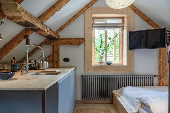 A compact kitchen with a wooden ceiling, a window with a view of greenery, a built-in TV, and a radiator.