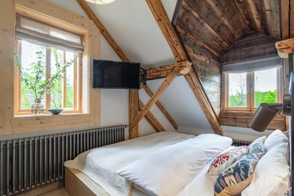 Un dormitorio con techo inclinado, vigas de madera, una cama con ropa blanca, un televisor montado y una ventana con vista a la vegetación.