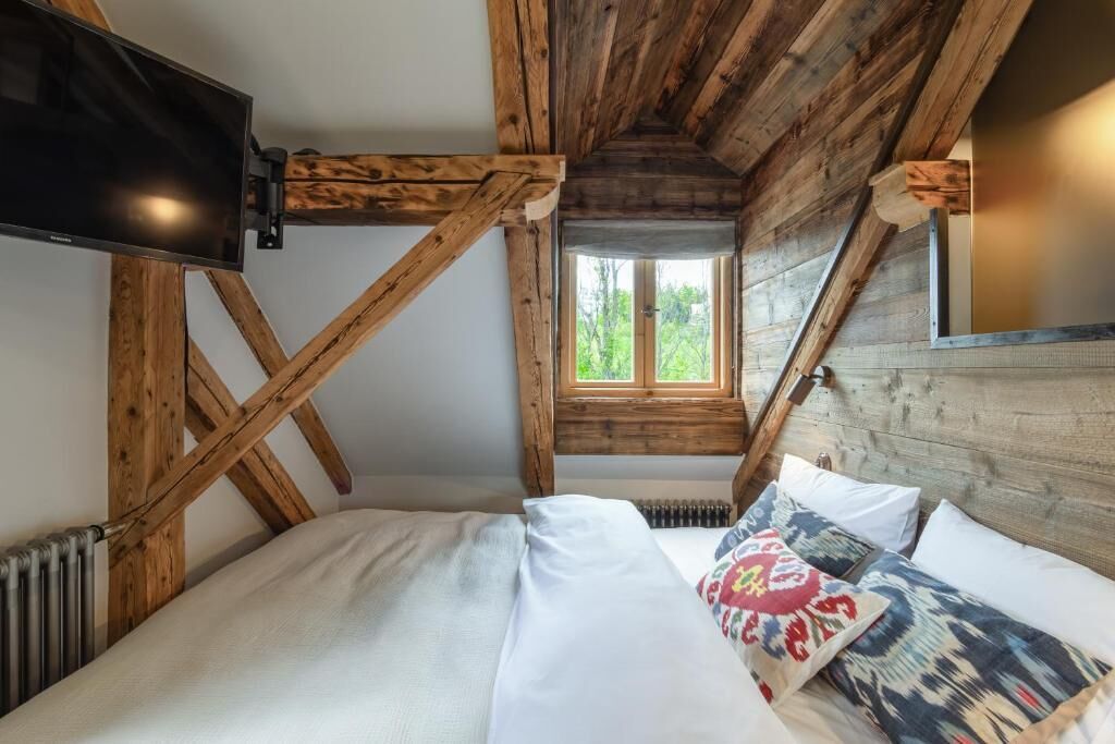 A bedroom with a wooden ceiling, a bed with white bedding, a TV mounted on the wall, and a window with a view of greenery.