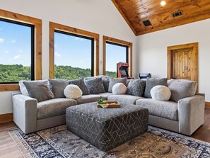 Ein modernes Wohnzimmer mit einem Ecksofa, einem Couchtisch und einem großen Fenster mit Blick auf einen Wald.