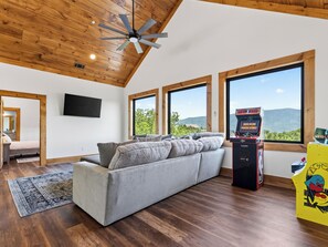 A modern living room with a gray sofa, wooden flooring, a ceiling fan, large windows, and a video game console. - Indoor Pool + Game Room + BBQ | Mins to Dollywood | Spurling Escape (Sevierville)