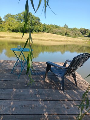 A wooden dock with a table and a chair by a calm lake, surrounded by trees. - Mobile Home "By the Lake" with Lake View and Private Garden (Naves)