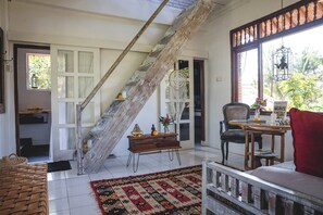 A room with a wooden staircase, a dining area with a table and chairs, a red sofa, and a view of greenery outside. - Brahma 11.11 Penestanan Ubud Apartment (Gianyar)