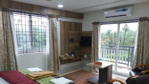 A hotel room with a bed, a sofa, a small table, a TV, and a balcony with a view of greenery. - Grand Meridian By Go2 Hotels and Resorts (Ambur)