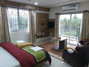 A hotel room with a bed, a sofa, a small table, a TV, and a balcony with a view of greenery. - Grand Meridian By Go2 Hotels and Resorts (Ambur)