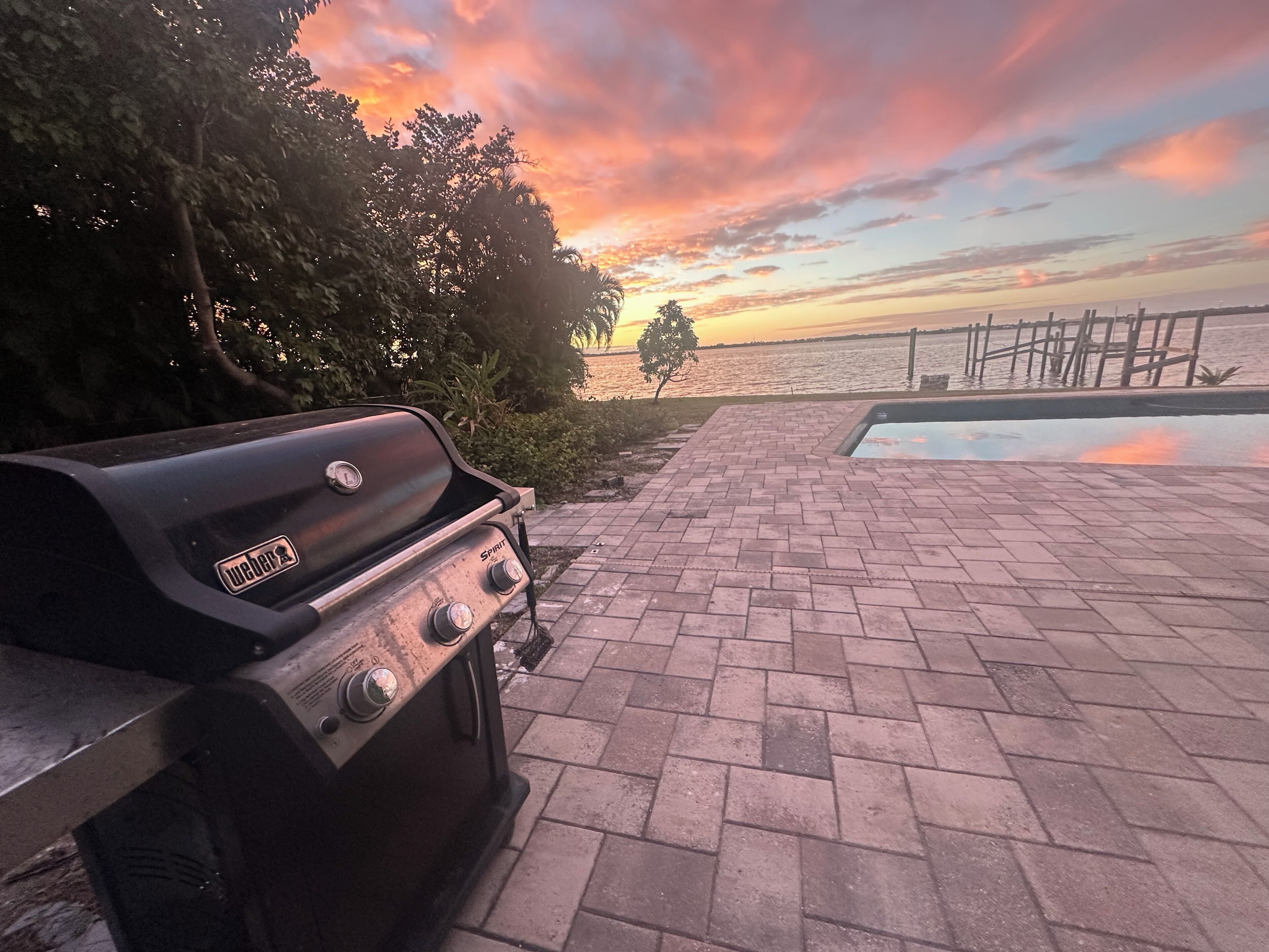 Una parrilla, un patio de ladrillo y vista al agua con un atardecer.