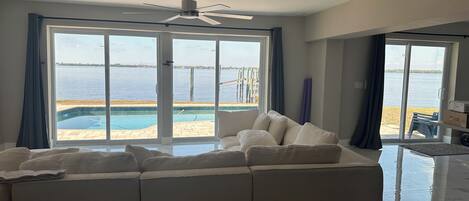 A modern living room with a sectional sofa, a ceiling fan, and large sliding glass doors leading to a pool and a view of the water.