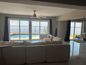 A modern living room with a sectional sofa, a ceiling fan, and large sliding glass doors leading to a pool and a view of the water. - Waterfront getaway, relaxation done right here (Fort Myers)