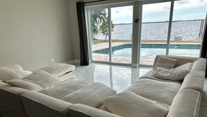 A spacious living area with a large beige sectional sofa, a glass sliding door leading to a pool, and a view of the water. - Waterfront getaway, relaxation done right here (Fort Myers)