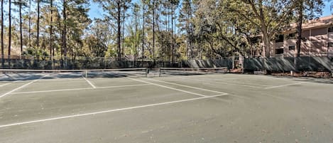 An outdoor tennis court surrounded by tall trees and a building in the background.