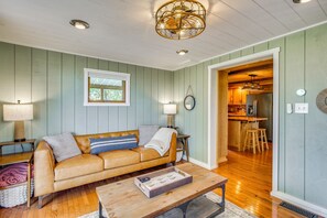 A living room with a sofa, coffee table, and lamps. There is a kitchen visible through an open doorway.