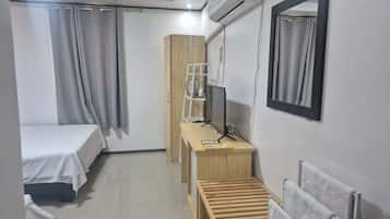 A hotel room with a bed, a desk with a computer, a radiator, and a window with curtains.