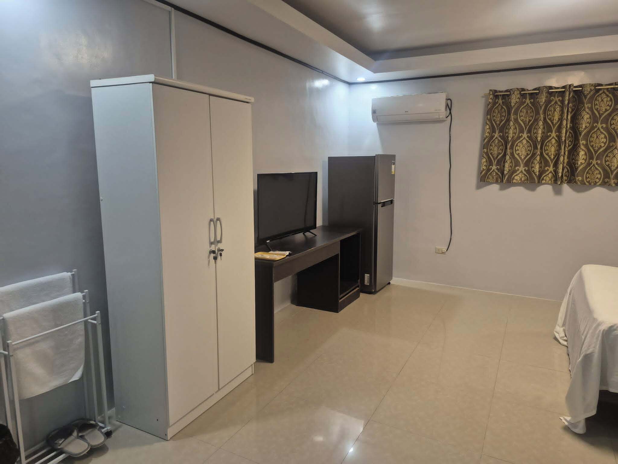 A small, clean room with a wardrobe, a desk with a computer, a bed, and a window with curtains.