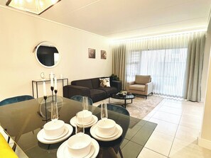 Ein modernes Wohnzimmer mit Esstisch für vier Personen, einer Couch, einem Couchtisch und einem großen Fenster mit Vorhängen.