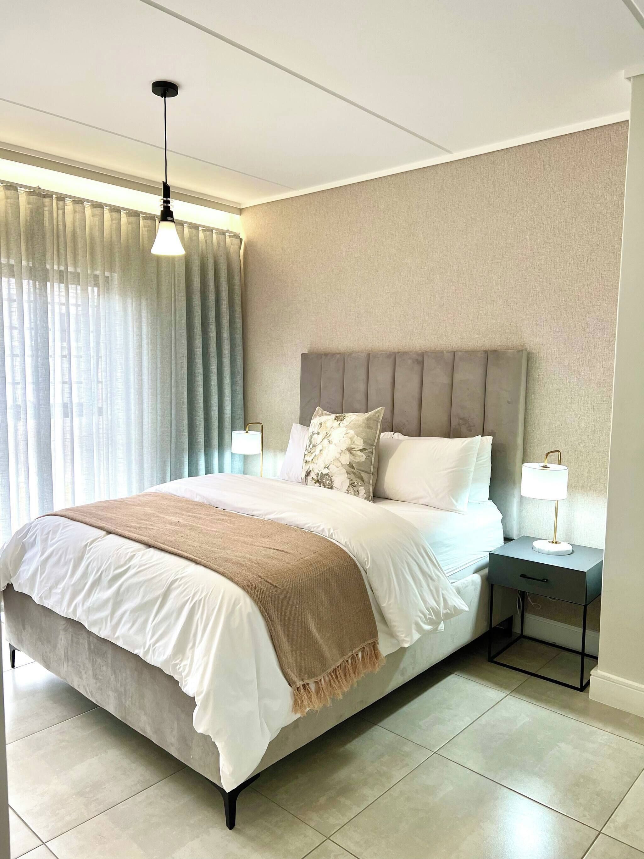A neatly made bed with a headboard, a bedside lamp, and a window with curtains.