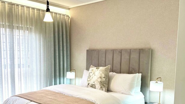 A neatly made bed with a headboard, a bedside lamp, and a window with curtains. - Luxurious 3-bedroom Apartment in Johannesburg's Waterfall Area (Johannesburg)
