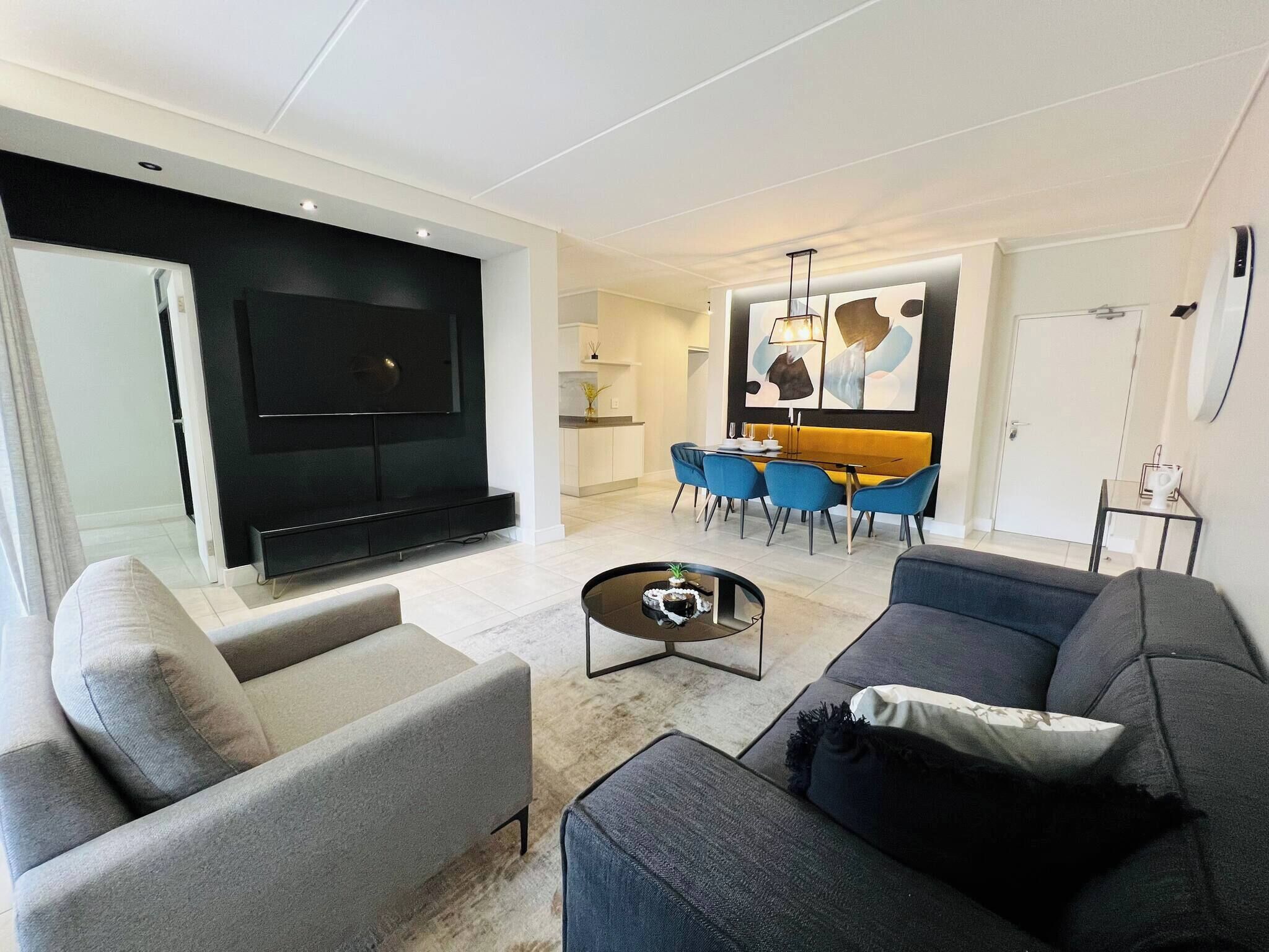 A modern living room with a flat-screen TV, a sofa, a coffee table, and a dining area with chairs and a table.