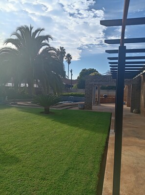 A well-maintained outdoor area with a grassy lawn, palm trees, and a brick structure. - Luxurious 4-Bedroom House Rental with Pool Access in Ngaka Modiri Molema (Ngaka Modiri Molema District Municipality)