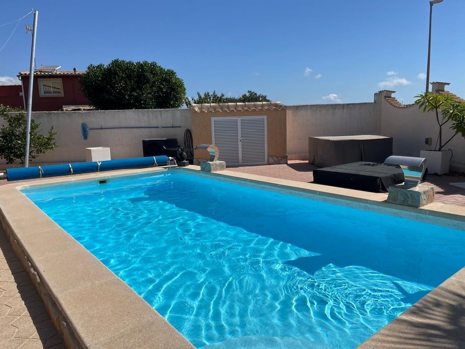 A clear blue swimming pool with a tiled deck, a covered seating area, and a wall-mounted air conditioning unit.