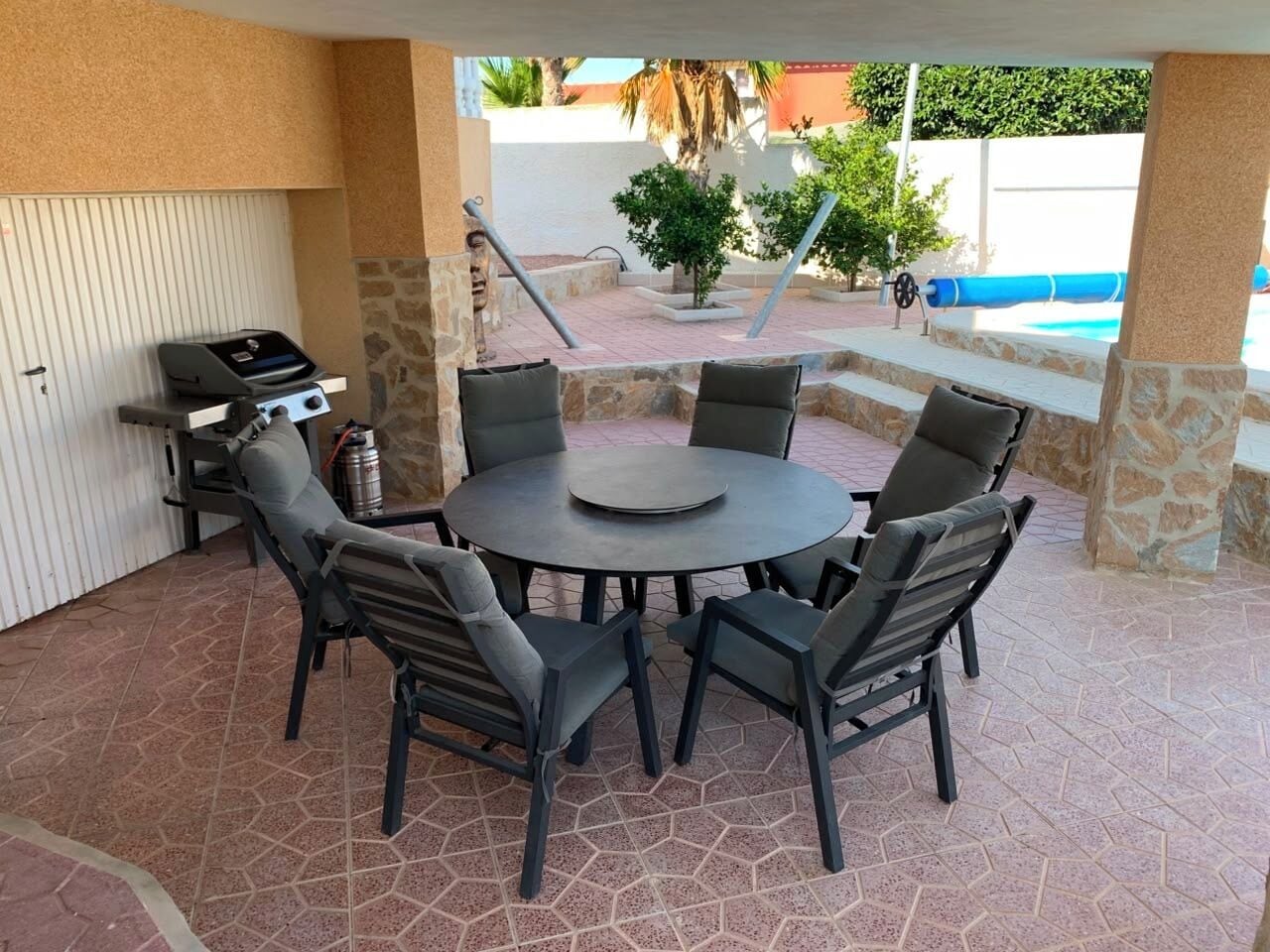 An outdoor patio with a round table and chairs, a grill, and a pool in the background.