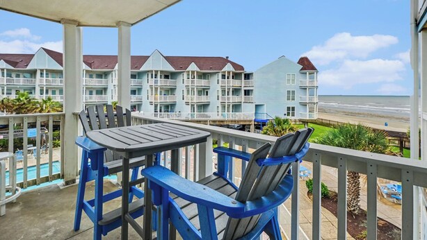 Ein Balkon mit Tisch und Stühlen, der Blick auf einen Strand und eine Reihe von Gebäuden mit Balkonen freigibt.