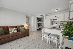 Ein kompakter Wohnbereich mit braunem Sofa, einer Essecke mit weißem Tisch und zwei Hocker sowie einer Küchenzeile mit weißen Schränken und Geräten.