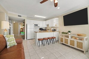 A modern kitchen with white cabinets, a central island with stools, and a flat-screen TV mounted on the wall. - Seascape Beachfront Condo w/Pool & Beach Views (Galveston)