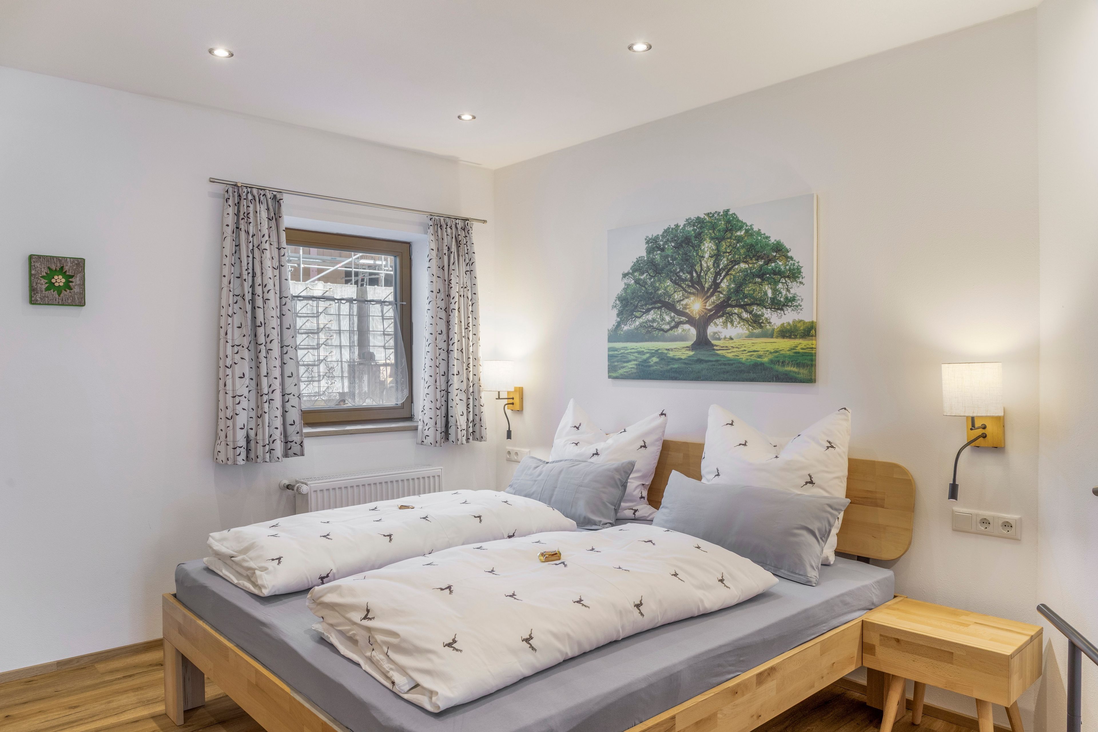 A bedroom with a bed, two bedside tables, a window with curtains, a radiator, and a wall-mounted painting.
