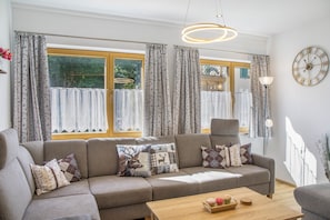 Ein modernes Wohnzimmer mit einem grauen Sofa, gemusterten Kissen, einem Holztisch mit Dekoration und einem großen Fenster mit Vorhängen.