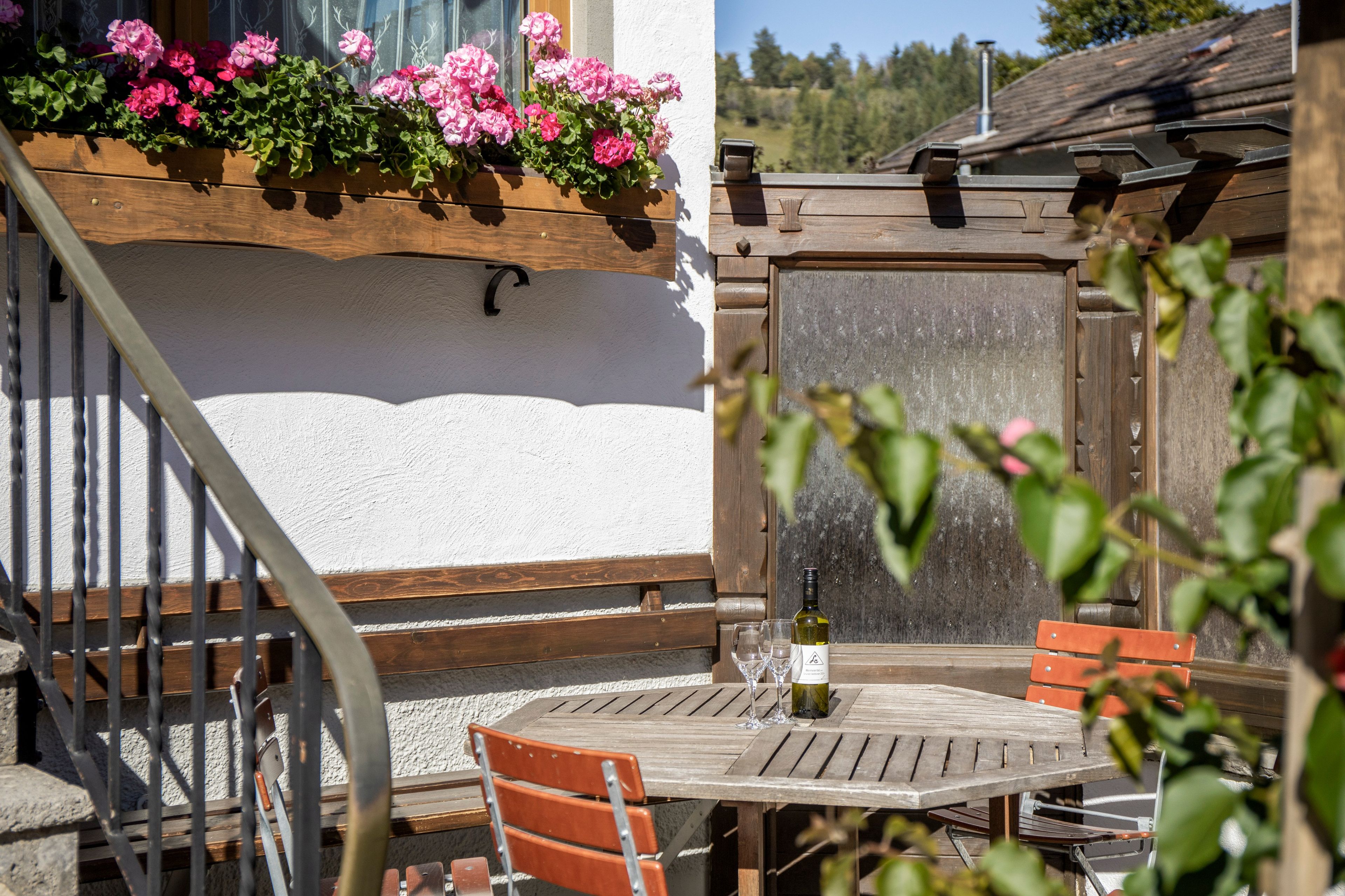 Ein hölzerner Sitzbereich im Freien mit Tisch und Stühlen, einer Weinflasche und einem Fenster mit rosa Blumen.