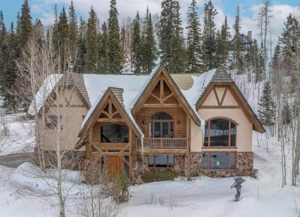 A log cabin with a stone foundation and wooden upper structure, surrounded by a snowy landscape and dense forest.