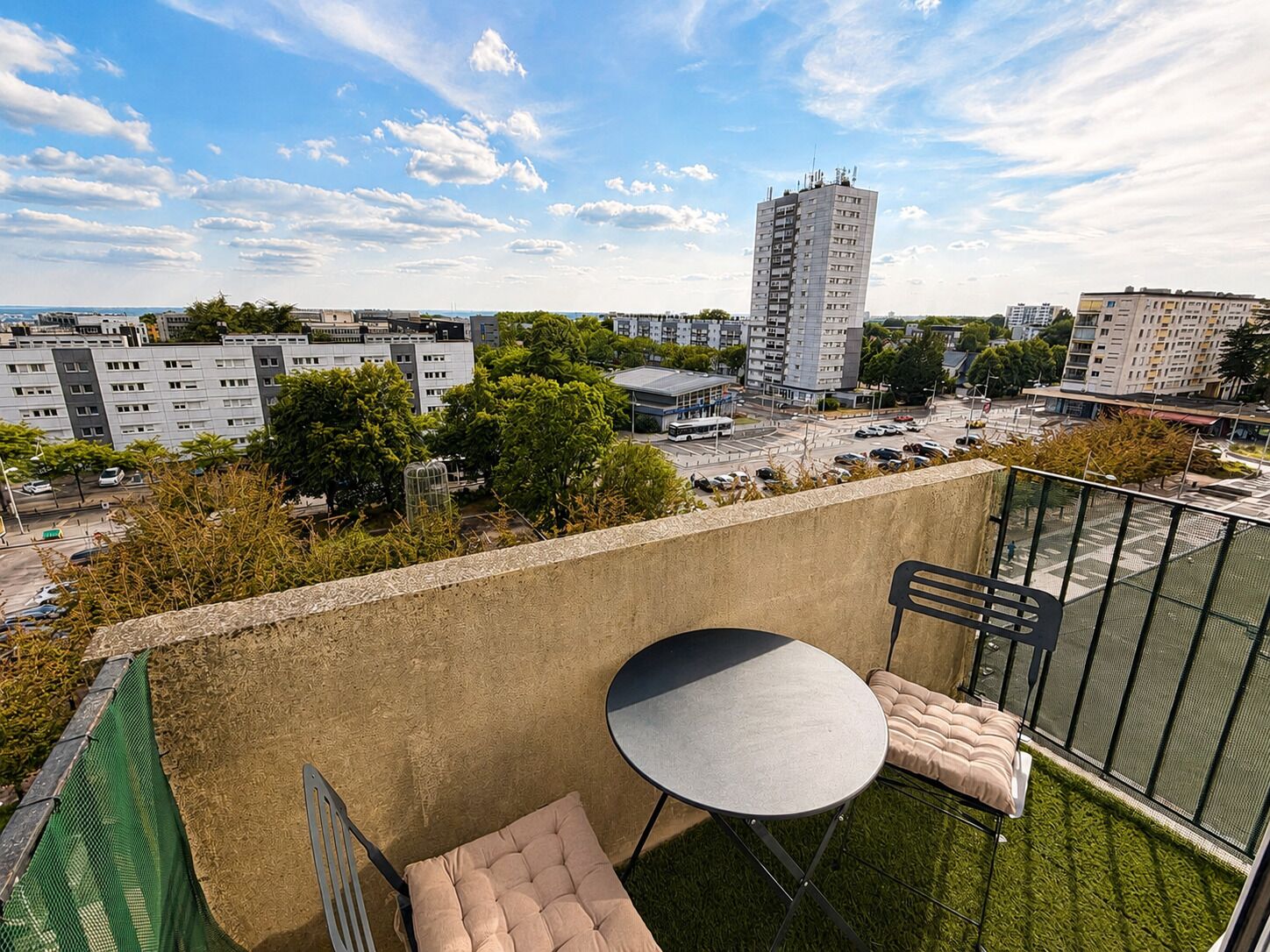 Un balcon avec une table et des chaises, donnant sur un paysage urbain composé d’immeubles et d’un parking.